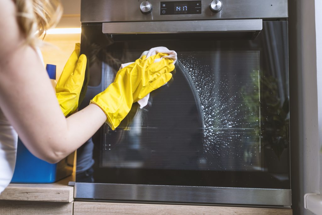 young smiling woman in protective glove with rag cleaning oven